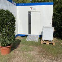 Zu sehen ist ein Wellblechcontainer mit einer Rampe, die zur Eingangstür führt- Direkt neben der Tür steht auf einer Holzpalette ein Kühlschrank.