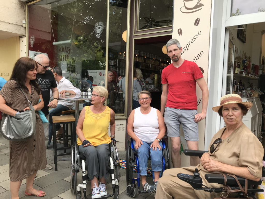 Das Bild zeigt Rita, zwei weitere Damen im Rollstuhl, Daniel und eine Journalistin vor der Caffee Bar.