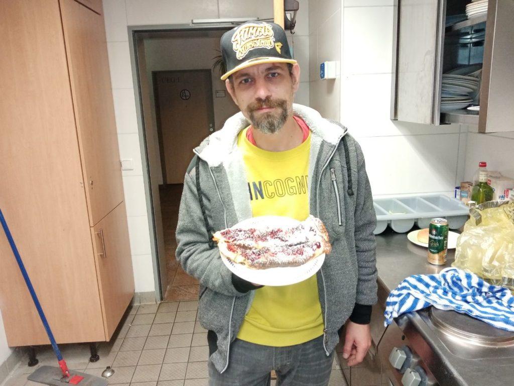 Hier steht ein Mann mit Basecap in einer Küche und präsentiert vor sich mehrere Pfannkuchen auf einem Teller.