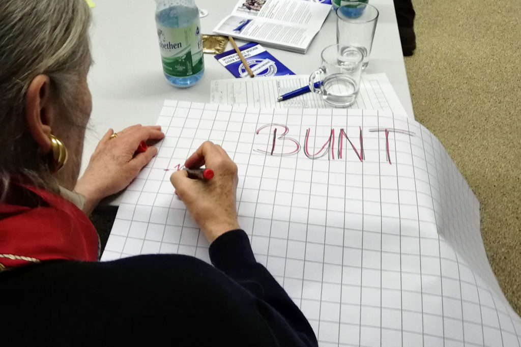 "Was ist bunt für mich?": Blick über die Schulter einer Teilnehmerin, die auf einen Flipchart gerade etwas zum Stichwort "BUNT" schreibt.