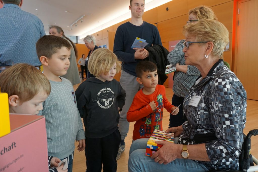 Rita (Frau im Rollstuhl rechts im Bild) erklärt 4 kleinen Kindern den Lego-Rampen-Bau