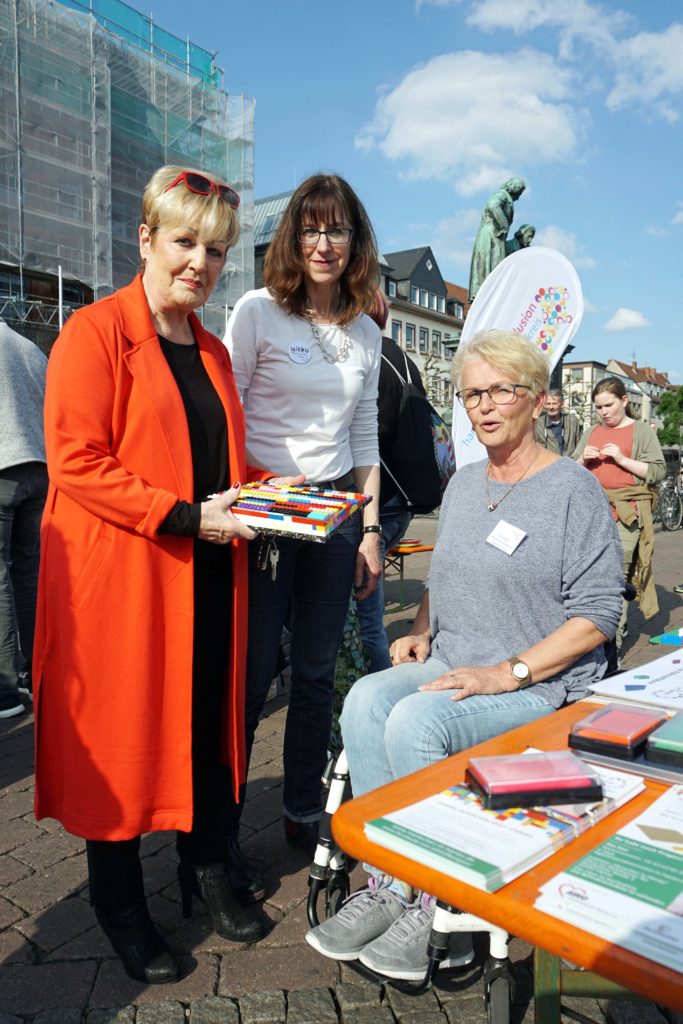 3 Frauen, von links in einem roten Mantel Frau Funck stehend, daneben Sylvie mit weißem T-shirt, daneben Rita mit grauem Pullover im Rollstuhl sitzend.