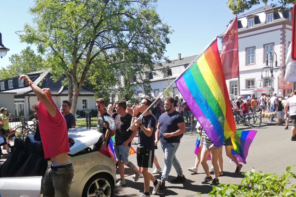 Wagen und Menschen teilen sich die Straße. Ein Teilnehmer hat auch eine Regenbogenflagge in der Hand.