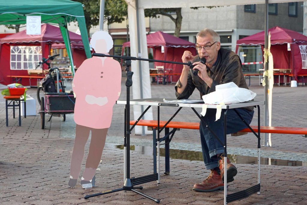 Jürgen Roth sitzt unter einem Zelt, vor ihm ein Mikrofon. Links daneben einer unserer Menschen in Hanau Pappfiguren.