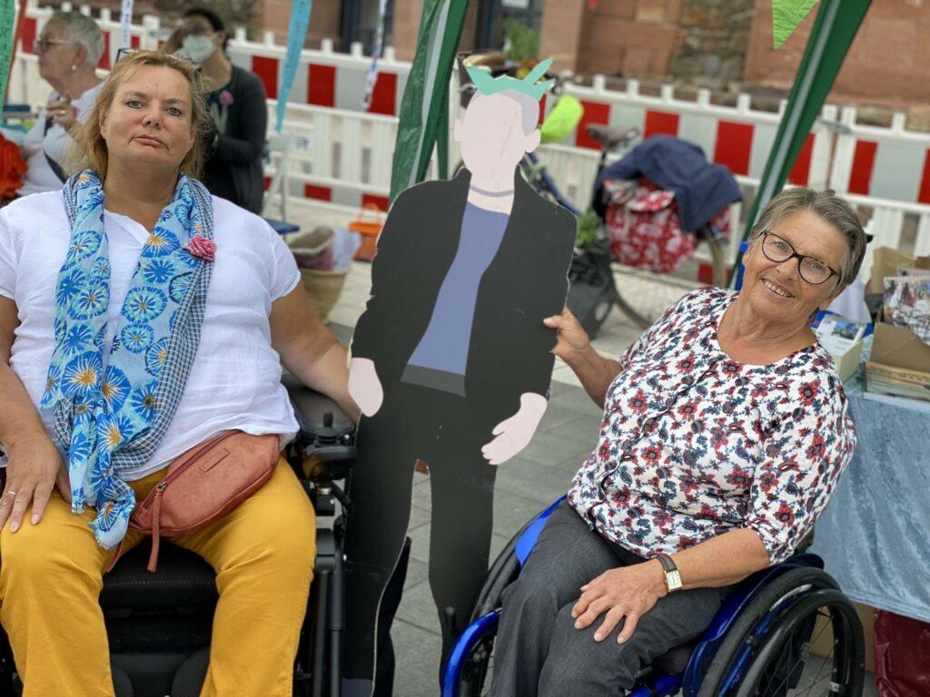 Kirsten und Marianne, beide im Rollstuhl, halten zwischen sich eine unserer Pappfiguren fest. Es ist der Punk.
