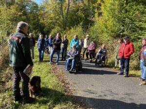 2021: Barrierefreie Waldführung. Viele Personen, zwei davon im Rollstuhl, sind auf einem asphaltierten Waldweg und hören dem Förster zu. Neben ihm ist sein Hund.