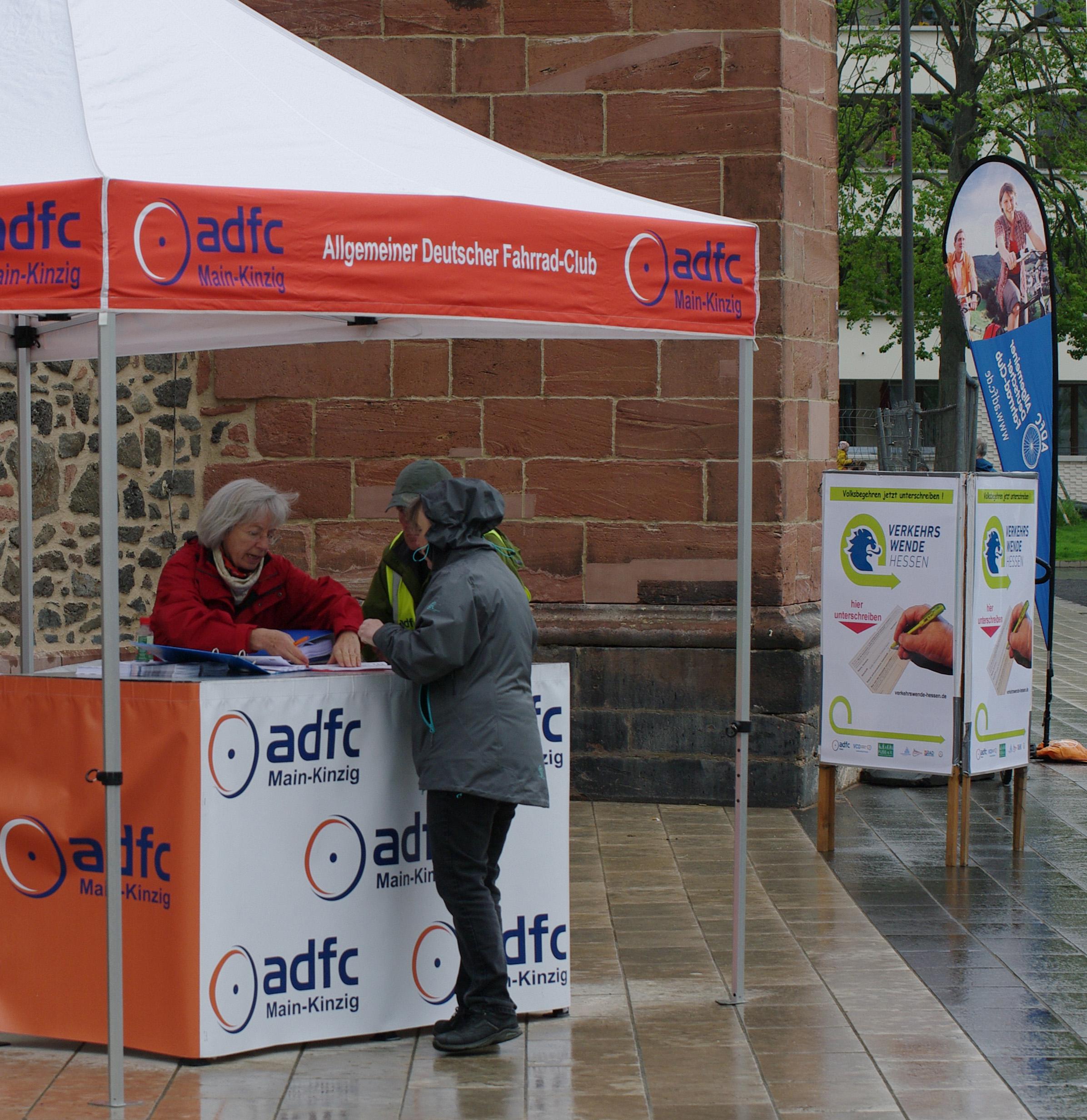 Ein Zelt mit dem Logo des adfc steht im Regen auf einem Platz . Drei Menschen stehen am Stand und sprechen mit einander.