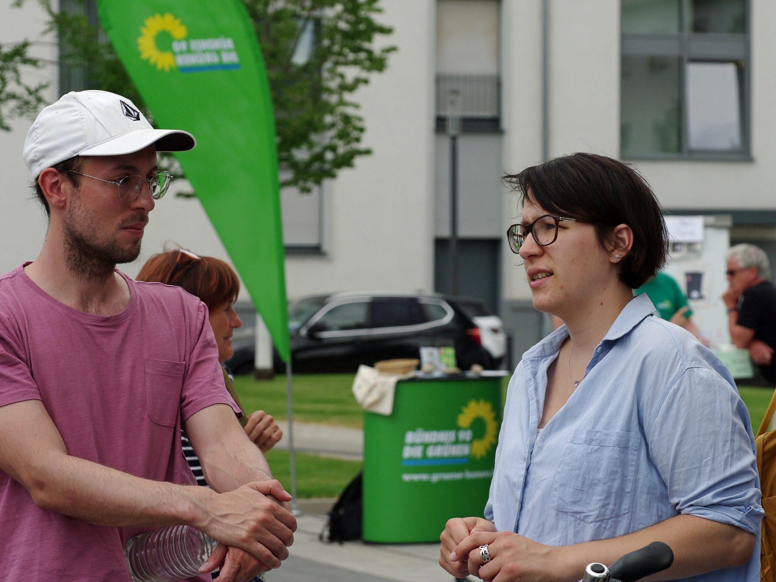 Eine Frau und ein Mann sind im Gespräch. Im Hintergrund ist die grüne Fahne von der Partei Bündnis 90/die Grünen zu sehen.