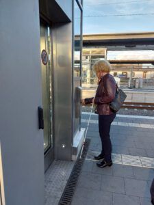 Eine Frau mit Langstock steht vor einem Aufzug auf dem Bahnsteig, und drückt den Knopf.