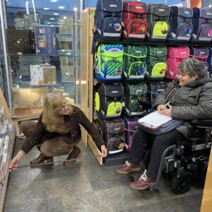 2023: Check eines Geschäftes. In einem Regal sind viele Schulranzen. Eine Frau im Rollstuhl hält Papier und Stift in den Händen und eine andere misst den Gang im Geschäft aus.