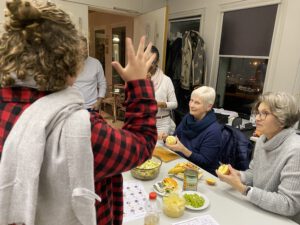 2024: Stilles Buffet. Eine junge Frau erklärt in Gebärdensprache. Sie steht vor einem Tisch mit Essen. Zwei Frauen sehen zu ihr und sind fröhlich.