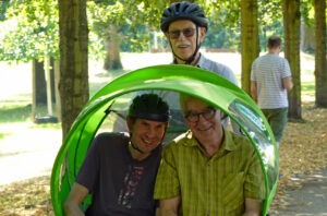 2 Männer in einer Rikscha mit einem Fahrer mit Helm und Sonnenbrille auf.