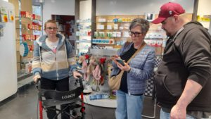 2025: Zwei Frauen, eine mit Rollator, und ein Mann sind in einer Apotheke und checken diese.