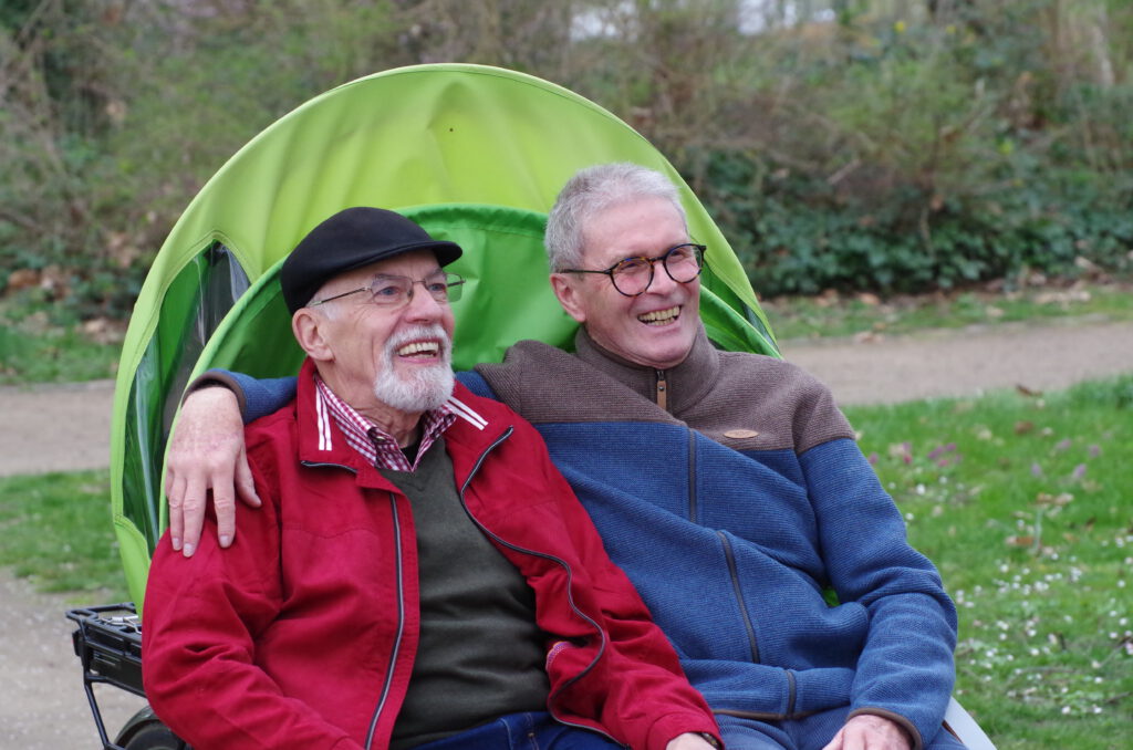 Ein Mann mit blauer und einer mit roter Jacke sitzen arm in arm in der RIkscha