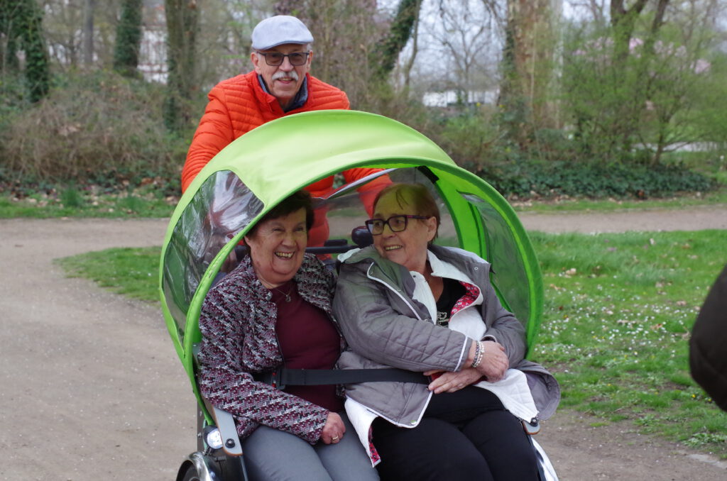 Zwei Frauen sitzen lachend und eingekuschelt in einer grünen Rikscha, dahinter fährt ein Pilot mit roter Jacke durch einen Park.