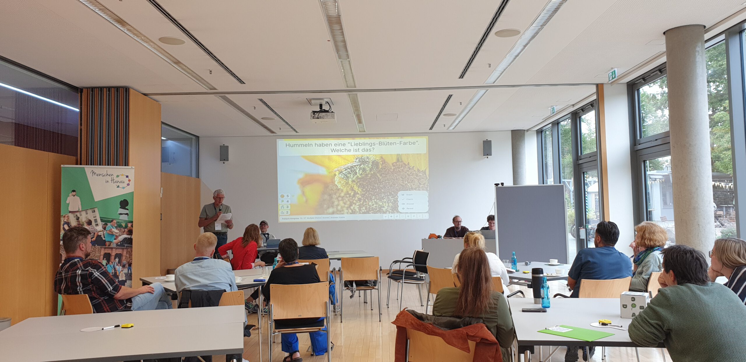 Foto vom Kongress „Dein Ehrenamt 2026“: Menschen sitzen in einem Veranstaltungsraum und hören einem Redner zu. Im Hintergrund ist eine Quizfrage über Hummeln an die Wand projiziert.