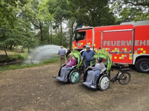 Zwei Rikschas mit Fahrgästen stehen auf einem Platz, daneben spritzt ein Feuerwehrauto eine Wasserfontäne.