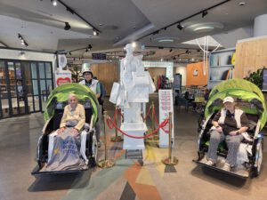 Zwei Rikschas mit Seniorinnen stehen in einer hellen Eingangshalle, in der Mitte eine große weiße Figur.