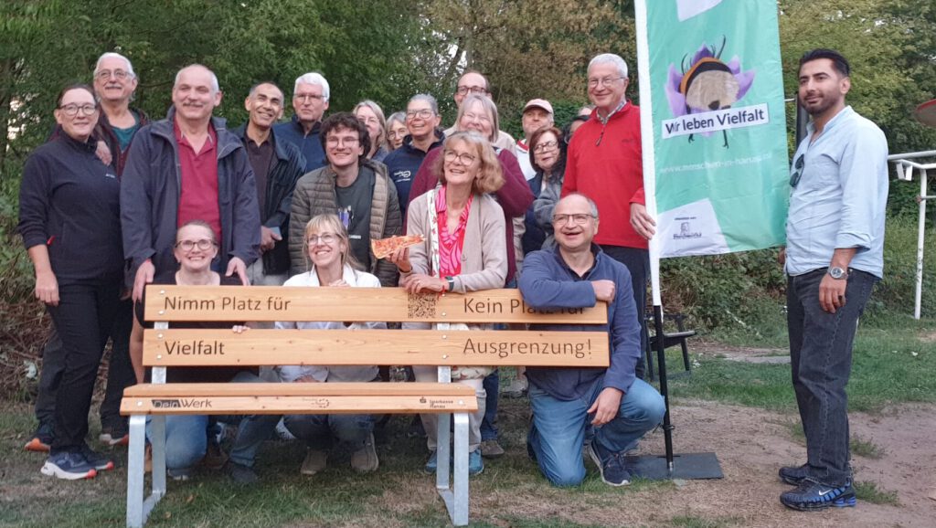 Foto vom Internationalen Beetgeflüster im Licht- und Luftbad Hanau: Gruppenfoto: Viele Teilnehmende stehen und sitzen rund um die neue Bank gegen Ausgrenzung, daneben die Vereinsfahne.