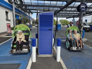Zwei Rikschas mit Fahrgästen stehen unter einem Vordach neben einer blauen Ladesäule.