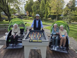 Zwei Rikschas mit Seniorinnen stehen an einem Tischkicker im Park, dazwischen steht ein Pilot mit Fahrradhelm.