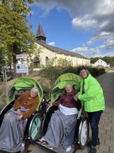 Ein Pilot in gelber Warnjacke steht neben zwei Rikschas mit Fahrgästen, im Hintergrund eine kleine Kirche unter blauem Himmel.