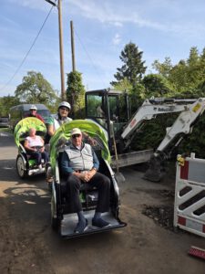 Zwei Rikschas mit Fahrgästen fahren an einer Baustelle mit einem kleinen Bagger vorbei.