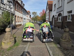 Zwei Rikschas mit Fahrgästen fahren nebeneinander durch eine Gasse mit Fachwerkhäusern.