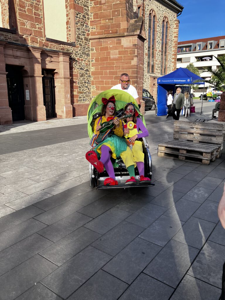 Eine Person im bunten Clownskostüm sitzt in einer Rikscha vor einer Kirche aus rotem Sandstein, dahinter ein Pavillon.