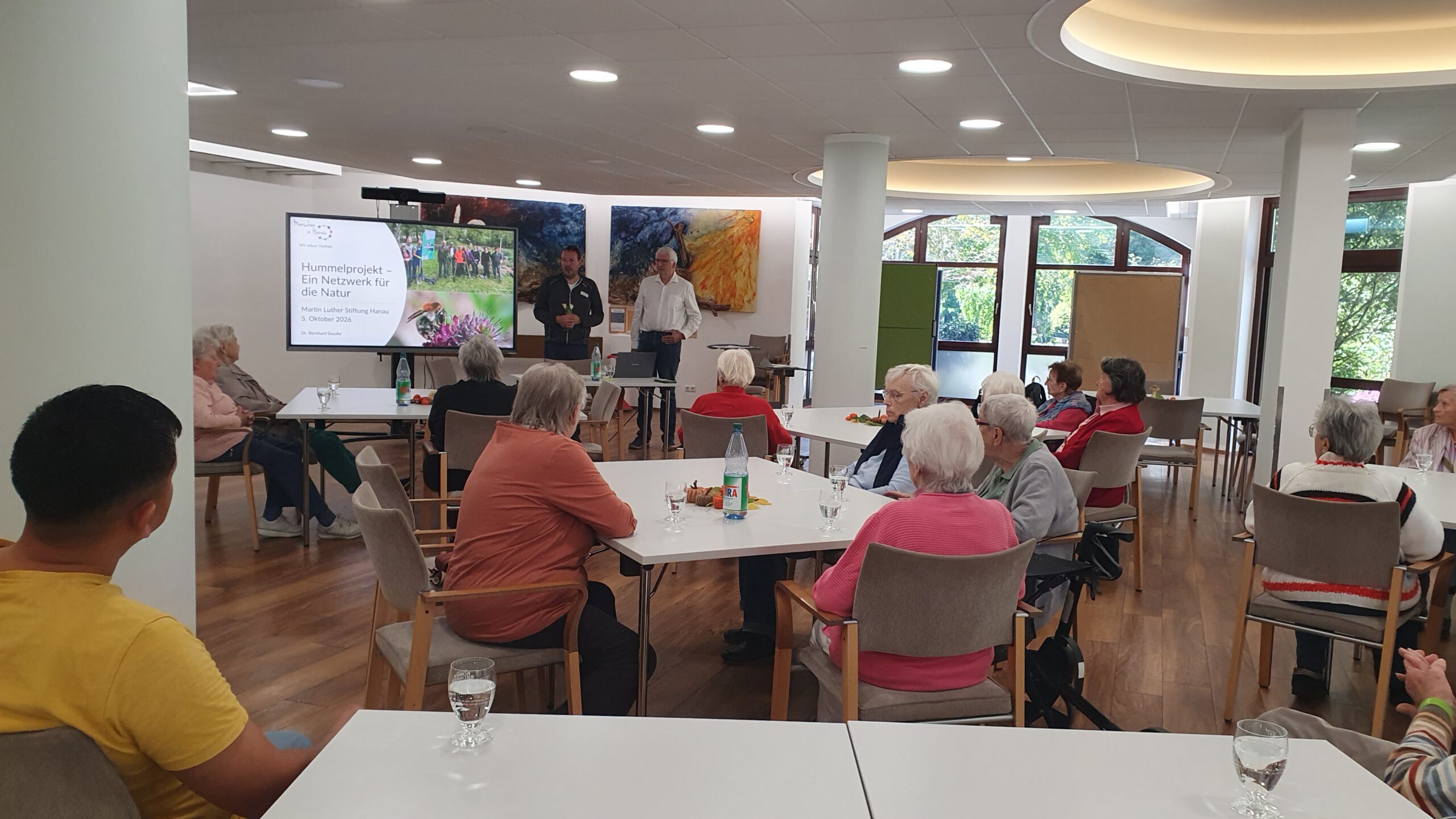Ältere Menschen sitzen an Tischen in einem hellen Gemeinschaftsraum und hören einem Vortrag zu, vorne ist eine Präsentation auf einem Bildschirm zu sehen.