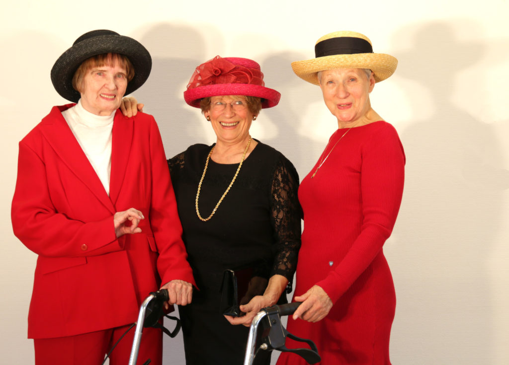 Drei ältere Frauen stehend mit Hut in roten und schwarzen Kleidern und Rollator.