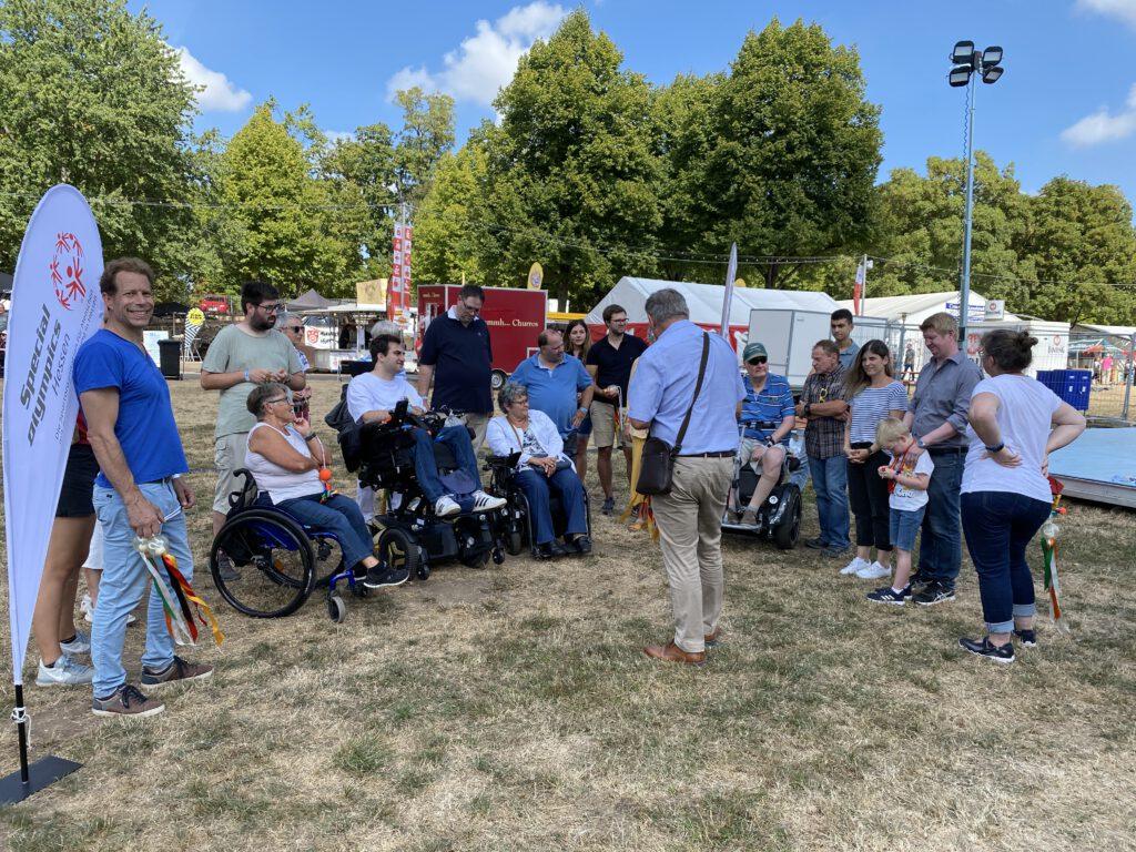 Oberbürgermeister Kaminsky spricht auf einer trockenen Wiese zu einer kleinen Gruppe aus Menschen, die zum Teil im Rollstuhl sitzen bzw. einen Blindenstock dabei haben.