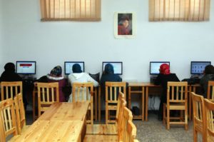 Das Bild zeigt einen großen Raum mit vielen Tischen. Am Ende des Raumes sitzen fünf Frauen vor Computern und lernen den Umgang mit ihnen.