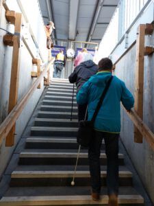 Zwei Personen mit Langstock steigen eine Treppe nach oben und halten sich am rechten Geländer fest.