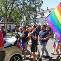 Teilnehmer des Events des Christopher Street Day laufen in Hanau, hinter einem Cabrio her. Sie tragen große Regenbogen-Flaagen und haben sie sich auch ins Gesicht gemalt. Im Hintergrund ist ein reges Treiben mit vielen verschiedenen Menschen, Fahrradfahrern und Hundebesitzern. Es sind alle Alter vertreten, von jung bis alt.