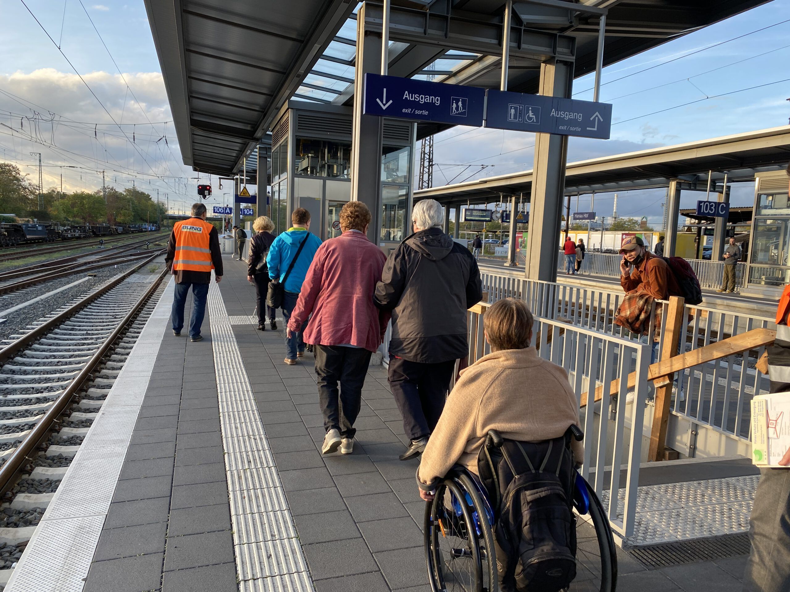 Eine Rollstuhlfahrerin und weitere Personen gehen einen Bahnsteig entlang. Weiße Leitstreifen heben sich von der grauen Bodenfliesen ab.