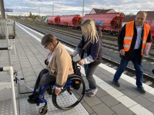 Eine Frau schiebt eine Rollstuhlfahrerin rückwärts aus dem Hublift auf einem Bahnsteig.