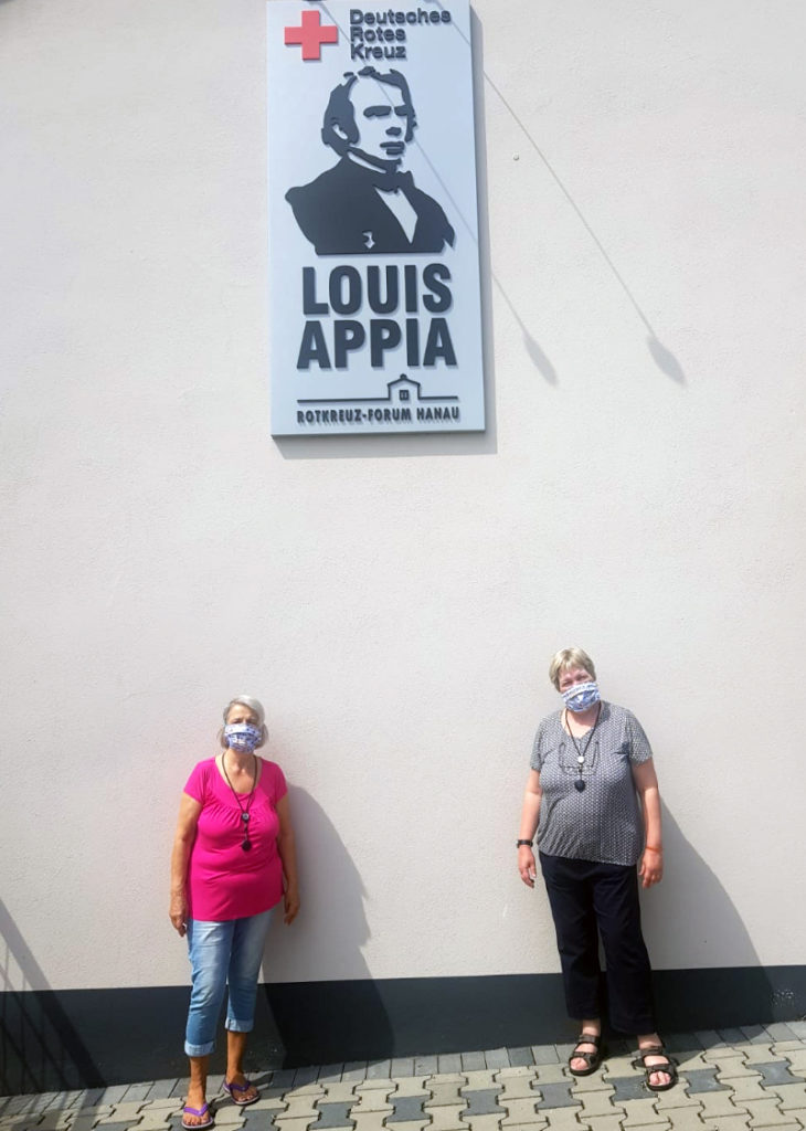 2 Frauen stehen vor einer Mauer mit Masken. Oben auf der Wand ist ein Logo, von Louis Appia DRK zu sehen.