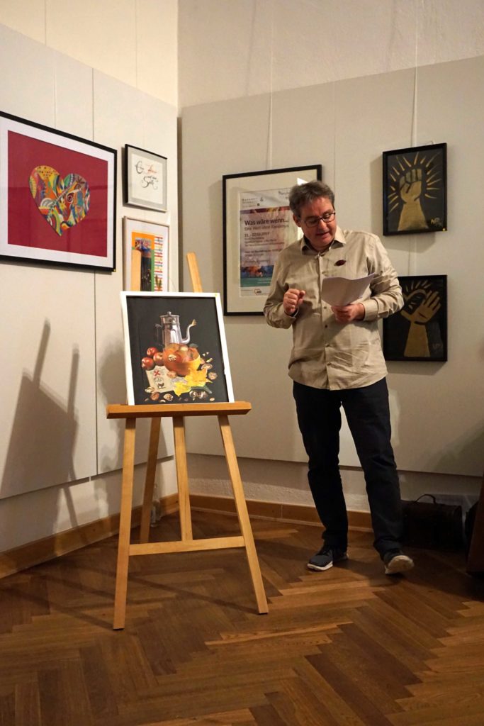 Man sieht Peter Koch stehend, wie er das Kunstwerk "Markttag" von Renate Mann beschreibt. Es steht links neben ihm auf einer Staffelei
