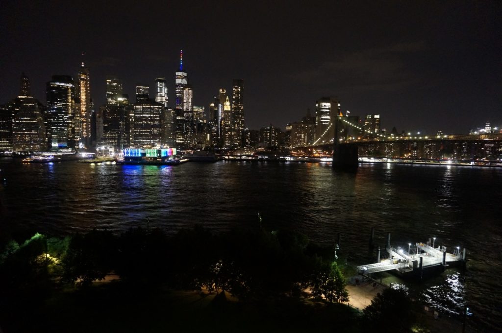 Blick auf Manhattan in der Nacht. Man sieht die Spitze vom One World Trade Center in Regenbogenfarben sowie ein Pier am East River.