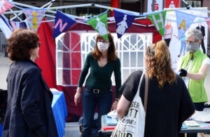 2020: Corona-Info-Stand: Zwei Frauen mit Stoffmasken stehen unter einem Stand von MiH auf dem Markplatz. Sie sprechen mit Passantinnen.