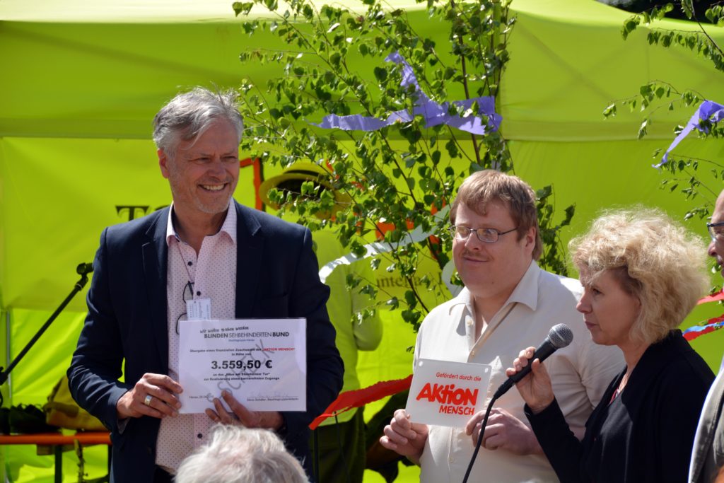 Silvia Schäfer übergibt einen Scheck der Aktions Mensch für den Bau der Rampe. Links neben ihr stehen Andreas Schild und Michael Stegmann den Scheck haltend.