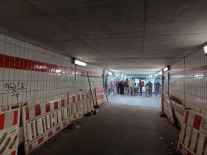 Ein langer dunkeler Fußgängertunnel ist im vorderen Drittel von rot-weissen Baustellenabsperrgittern eingegrenzt. Am Ende des Tunnels steht eine Gruppe von Menschen.
