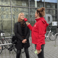 Sylvie, eine Frau in schwarzer Jacke und mit blonden Harren wird von der Reporterin in roter Jacke und mit rotem Mikrofon interviewt