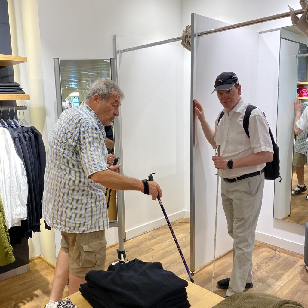 Ein Mann mit Taststock und ein Mann mit Krücken checken eine Umkleidekabine.