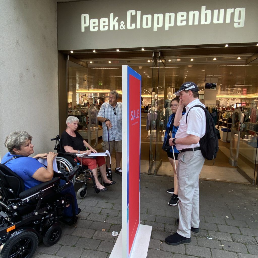 Zwei Frauen im Rollstuhl, ein Mann mit Taststock und ein Mann mit Krücken sind am Eingang der Peek & Cloppenburg-Filiale in Hanau.