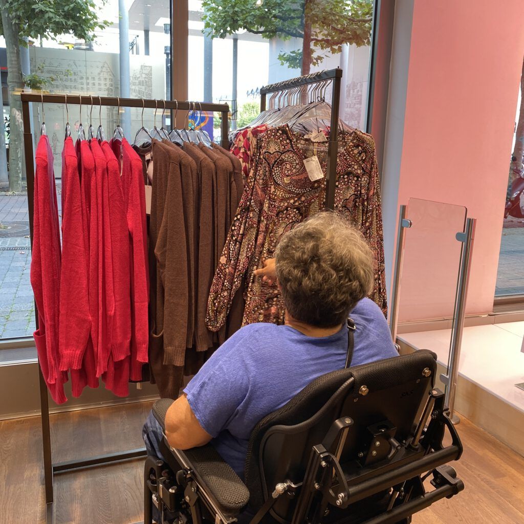 Eine Frau im Rollstuhl greift nach Kleidung, die an einem Kleiderständer hängt.