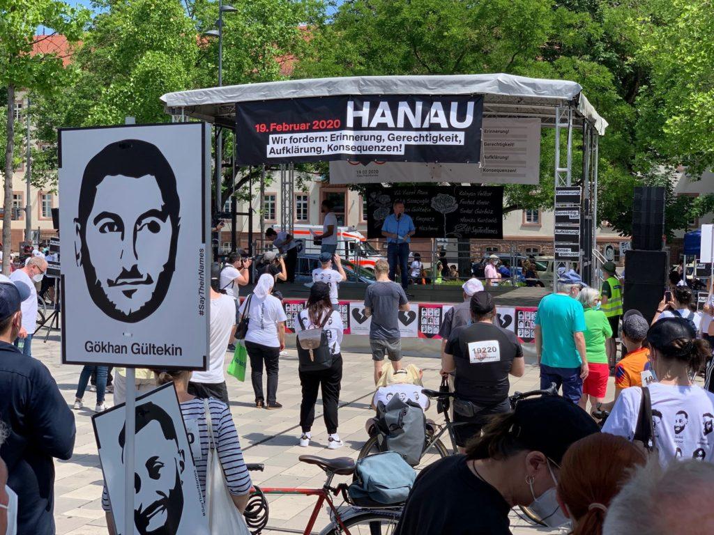 Ein Blick aus der versammelten Menschenmasse auf die Bühne. Im Vordergrund sieht man Menschen, die die Schilder mit den Namen der Opfer hochhalten. Im Hintergrund auf der Bühne ist der Oberbürgermeister Claus Kaminsky zu sehen.