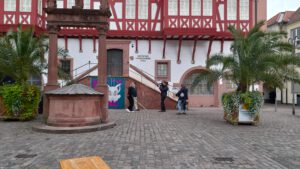 Boris und Kate Menzyk laufen auf dem Platz vor dem Goldschmiedehaus an der dortigen Treppe entlang. Ein Mann mit Kamera und ein Mann mit Mikrofon nehmen die beiden auf.