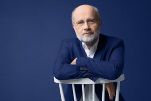 Mann mit Glatze weißem Bart und im blauen Anzug vor einer blauen Wand.